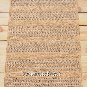 May include: Rectangular jute rug with a natural tan color and woven design. The rug features horizontal stripes with alternating tan and gray sections. The text "DurrieIndiaArt" is printed on the rug.