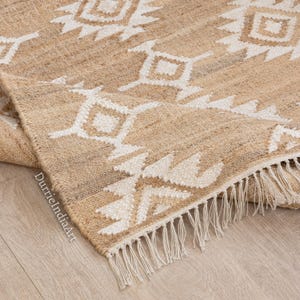 May include: A beige and white woven rug with a geometric pattern. The rug has a natural, textured appearance with a fringe along one edge. The design features repeating diamond and triangular shapes.