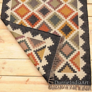 May include: A black, brown, and beige handwoven rug with a geometric pattern. The rug has a fringe border and is folded in half. The rug is on a wooden floor.