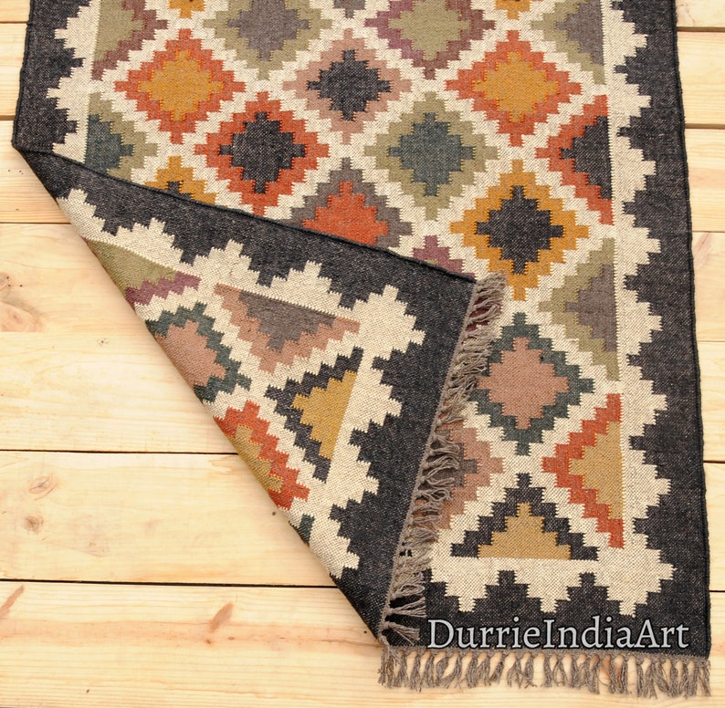May include: A black, brown, and beige handwoven rug with a geometric pattern. The rug has a fringe border and is folded in half. The rug is on a wooden floor.