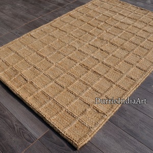 May include: A rectangular area rug made of natural tan woven fibres featuring a raised grid pattern of squares across the surface. The rug has a thick, textured border and includes the text DurrieIndiaArt printed in the lower right corner.