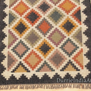 May include: A handwoven rug with a geometric pattern in shades of brown, black, orange, and beige. The rug has a black border and fringe.