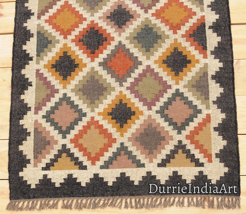 May include: A handwoven rug with a geometric pattern in shades of brown, black, orange, and beige. The rug has a black border and fringe.
