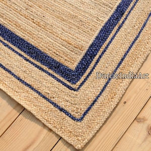 Könnte beinhalten: Ein rechteckiger Jute-Teppich in Naturbeige mit einem marineblauen Rand. Der Teppich hat eine gewebte Textur und liegt auf einer Holzoberfläche. Der Text "DurrieIndiaArt" ist sichtbar.