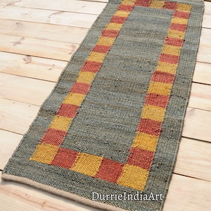 Puede incluir: Una alfombra rectangular gris y marrón con un borde a cuadros rojos y amarillos. La alfombra está hecha de fibras naturales y tiene un aspecto rústico.
