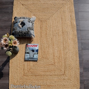 Può includere: Un tappeto rettangolare in juta con un design intrecciato. Un cuscino a fantasia blu e bianco, un vaso di fiori e una rivista con la parola "cover" sono sul tappeto. Il tappeto è su un pavimento in legno scuro.