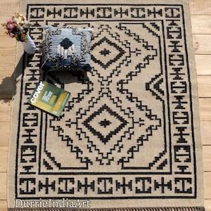 Puede incluir: Una alfombra beige y negra con estampado geom&eacute;trico y flecos. La alfombra tiene un coj&iacute;n blanco y negro encima. El texto "DurrieIndiaArt" es visible en la esquina inferior izquierda de la alfombra.
