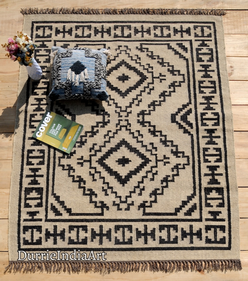 Puede incluir: Una alfombra beige y negra con estampado geom&eacute;trico y flecos. La alfombra tiene un coj&iacute;n blanco y negro encima. El texto "DurrieIndiaArt" es visible en la esquina inferior izquierda de la alfombra.
