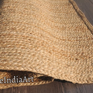 K&ouml;nnte beinhalten: Ein Naturfaser-Teppich aus Jute, einem nachhaltigen und strapazierf&auml;higen Material. Der Teppich hat eine gewebte Textur und eine nat&uuml;rliche beige Farbe. Der Teppich ist aufgerollt und die Kante ist sichtbar. Der Text "DurrieIndiaArt" ist auf dem Teppich sichtbar.