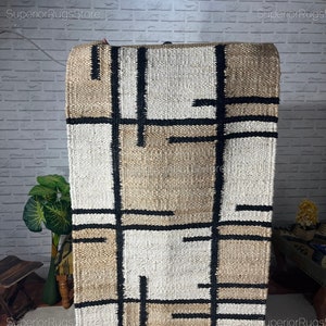Puede incluir: Una alfombra de yute con estampado geométrico con rayas negras, blancas y beige. La alfombra está hecha de fibras naturales y tiene una textura tejida a mano.