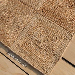 Puede incluir: Una alfombra de fibra natural hecha de yute, con un patrón tejido de cuadrados. La alfombra es de color marrón claro y tiene una textura rugosa.