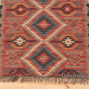 May include: A red, blue, and beige patterned rug with a geometric design. The rug has fringe on the bottom and the text "DurrieIndiaArt" is visible in the bottom right corner.
