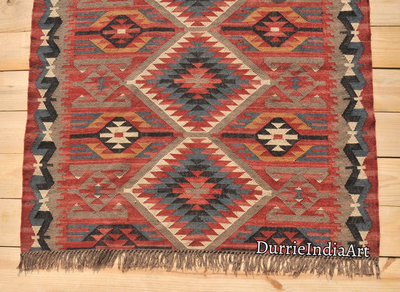 May include: A red, blue, and beige patterned rug with a geometric design. The rug has fringe on the bottom and the text "DurrieIndiaArt" is visible in the bottom right corner.
