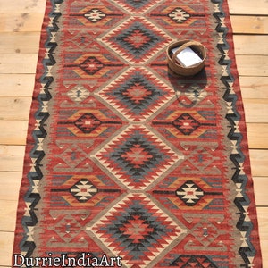 May include: A red, blue, and beige patterned rug with a geometric design. The rug has fringe on the edges and is made of wool.