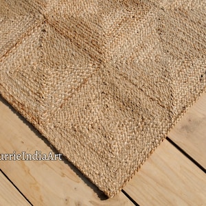 May include: A natural fiber rug with a geometric diamond pattern. The rug is made of woven jute in shades of tan and brown. The rug's texture is visible, and the corner of the rug is shown on a wooden surface.