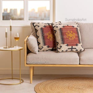 Puede incluir: Dos cojines cuadrados con un diseño geométrico en rojo, negro y dorado descansan sobre un sofá beige. Los cojines tienen una apariencia tejida y texturizada. Una pequeña mesa auxiliar redonda con tapa de mármol y patas doradas sostiene una copa de vino y dos candelabros.