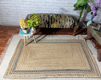May include: A rectangular jute rug with a black and white border. The rug is woven with a natural fiber and has a subtle texture. The rug is placed on a light brown wooden floor.