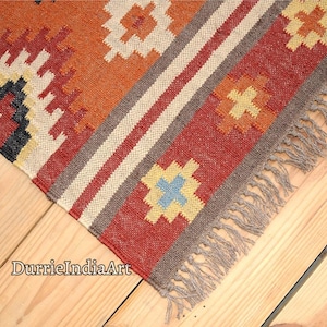 May include: A close-up shot of a vibrant woven rug with geometric patterns. The rug displays a mix of red, orange, cream, and grey hues. The design incorporates stripes and cross-like motifs. The rug has a fringed edge and is placed on a wooden surface.