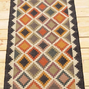 May include: A rectangular jute rug with a geometric pattern in shades of brown, orange, and beige. The rug has a black border and fringe on the edges. The text "DurrieIndiaArt" is printed on the bottom of the rug.