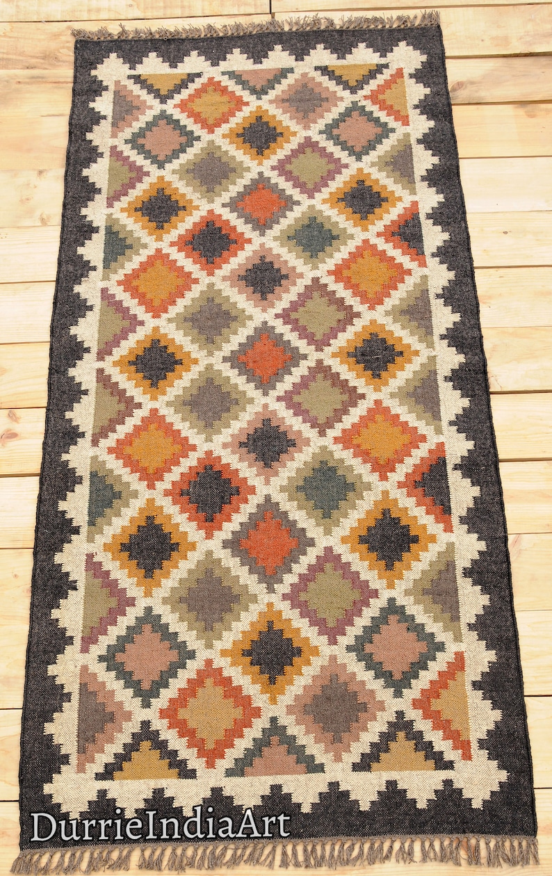 May include: A rectangular jute rug with a geometric pattern in shades of brown, orange, and beige. The rug has a black border and fringe on the edges. The text "DurrieIndiaArt" is printed on the bottom of the rug.