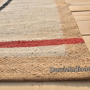 May include: A handwoven jute rug with a white background and black and red stripes. The rug is made of natural fibers and has a rustic look. The text "DurrieIndiaArt" is visible in the lower right corner of the rug.