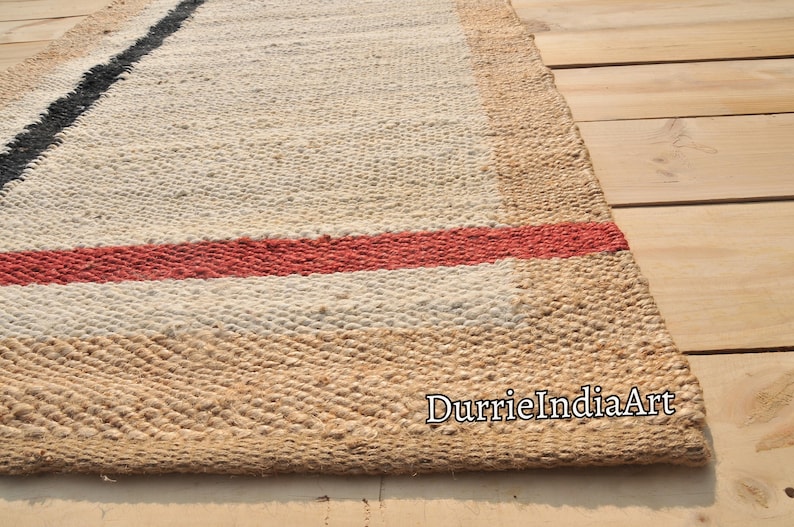 May include: A handwoven jute rug with a white background and black and red stripes. The rug is made of natural fibers and has a rustic look. The text "DurrieIndiaArt" is visible in the lower right corner of the rug.