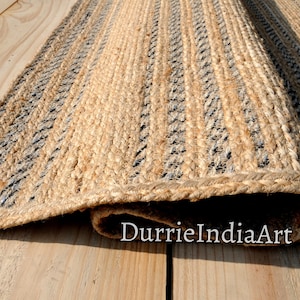 May include: A rectangular jute rug with a natural tan color and vertical stripes in black and gray. The rug is rolled up at the bottom, revealing its woven texture. The rug is placed on a light-colored wooden surface.