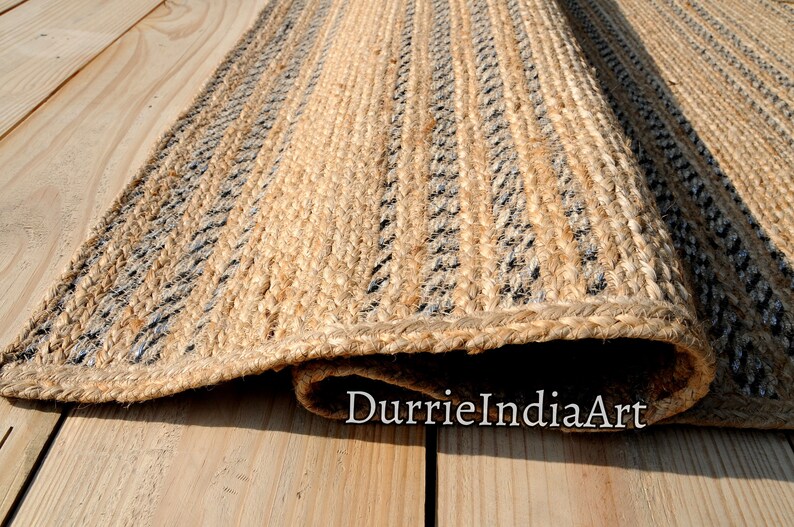 May include: A rectangular jute rug with a natural tan color and vertical stripes in black and gray. The rug is rolled up at the bottom, revealing its woven texture. The rug is placed on a light-colored wooden surface.