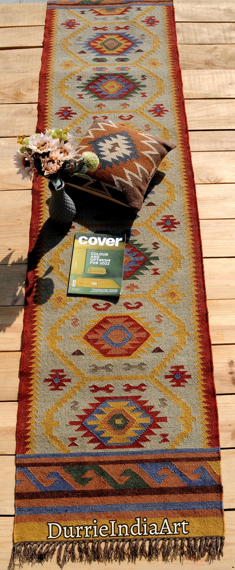Pu&ograve; includere: Un tappeto colorato con motivi geometrici e frange alle estremit&agrave;. Il tappeto &egrave; steso su un pavimento di legno con un cuscino decorativo e una rivista sopra. La copertina della rivista dice "Cover" e "Colore e ottimismo per il 2022". Il tappeto &egrave; fatto di juta e lana e ha un design tradizionale.
