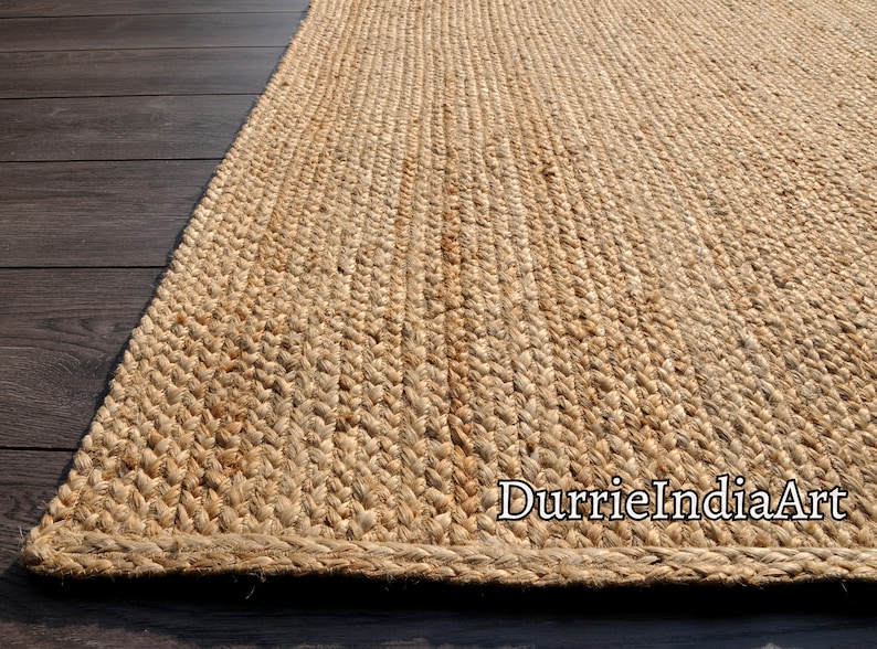 Pode incluir: Um tapete de fibra natural feito de juta, um material sustent&aacute;vel e dur&aacute;vel. O tapete tem uma textura tecida e uma cor bege natural. O tapete &eacute; mostrado em um piso de madeira. O texto "DurrieIndiaArt" &eacute; vis&iacute;vel no canto inferior direito da imagem.