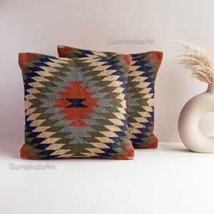 Puede incluir: Dos cojines decorativos con un patrón geométrico en tonos azules, verdes, beige y naranja. Los cojines están hechos de tejido tejido y tienen una superficie texturizada.