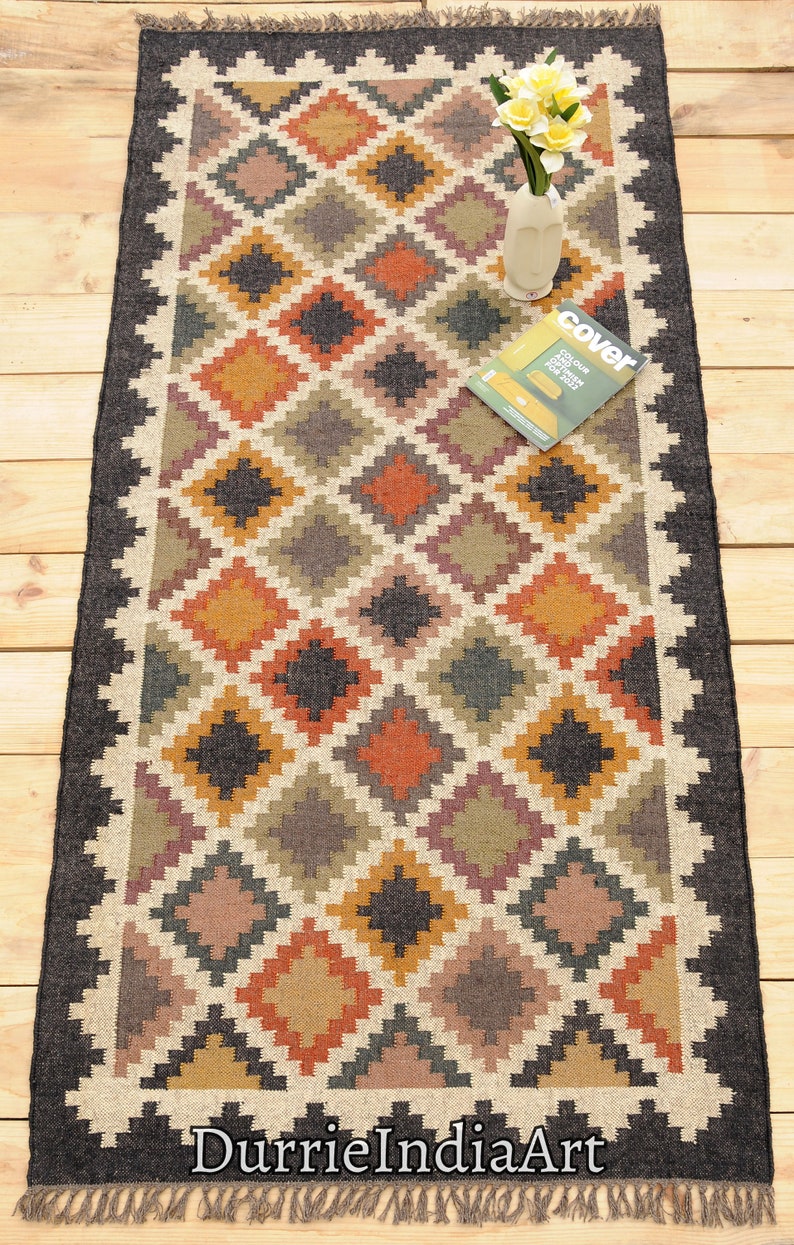 May include: A rectangular jute rug with a geometric pattern in shades of brown, orange, yellow, and black. The rug has a black border and fringe on the edges. The text "DurrieIndiaArt" is printed on the rug.