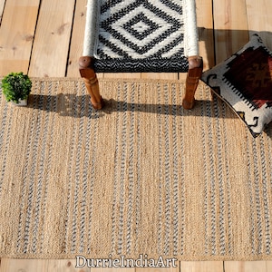 May include: A rectangular jute rug with vertical stripes in natural and black tones. The rug is placed on a wooden surface, with a chair, a decorative pillow, and a small potted plant nearby. The rug's texture and design create a rustic aesthetic.