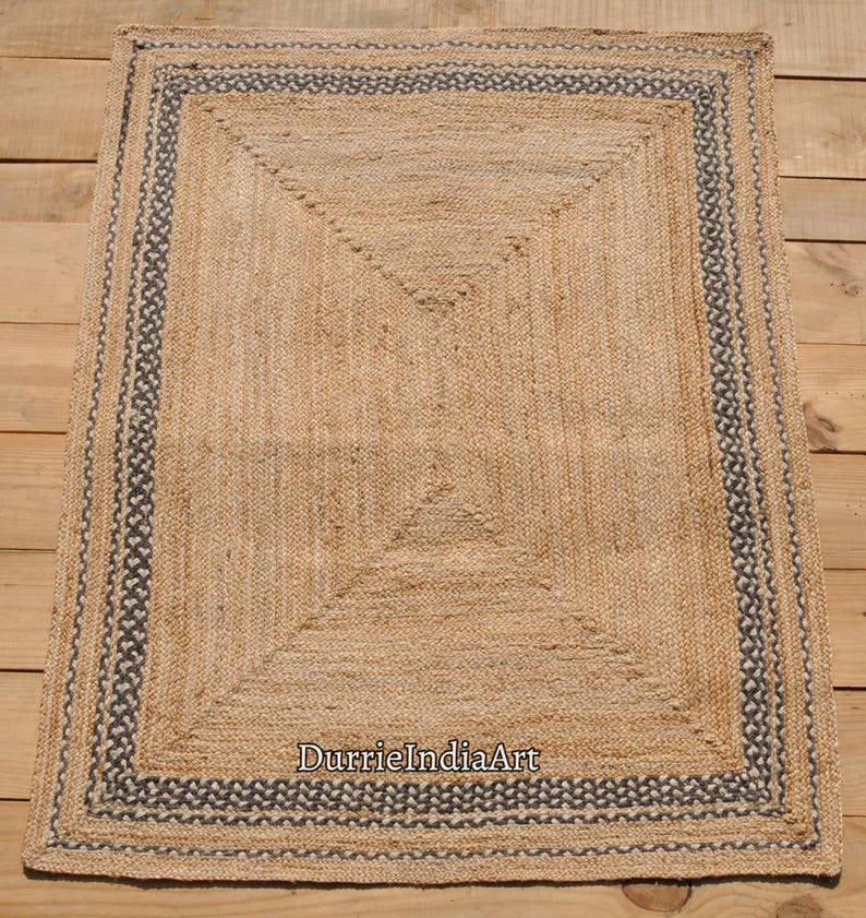 Puede incluir: Una alfombra de fibra natural con un dise&ntilde;o tejido y un borde gris. La alfombra est&aacute; hecha de yute y tiene un color natural y terroso.