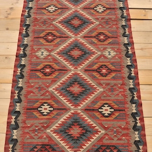 May include: A red, blue, and beige handwoven rug with a geometric pattern. The rug has a fringe border and is made of wool.
