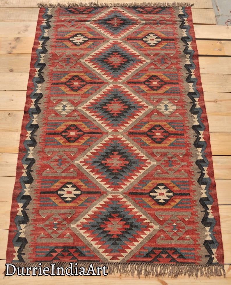 May include: A red, blue, and beige handwoven rug with a geometric pattern. The rug has a fringe border and is made of wool.