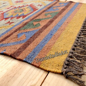 Pu&ograve; includere: Un tappeto colorato tessuto a mano con un motivo geometrico in tonalit&agrave; di marrone, blu, giallo e rosso. Il tappeto ha un bordo a frangia ed &egrave; realizzato in fibre naturali.