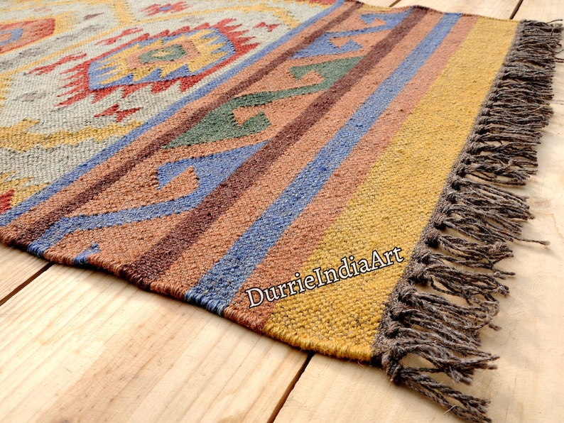 Pu&ograve; includere: Un tappeto colorato tessuto a mano con un motivo geometrico in tonalit&agrave; di marrone, blu, giallo e rosso. Il tappeto ha un bordo a frangia ed &egrave; realizzato in fibre naturali.