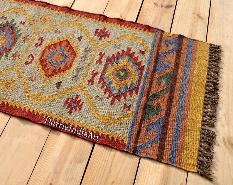 Pu&ograve; includere: Un tappeto colorato tessuto a mano con motivi geometrici in tonalit&agrave; di rosso, giallo, blu e verde. Il tappeto ha frange alle estremit&agrave; ed &egrave; fatto di lana.