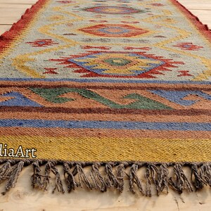 Pu&ograve; includere: Un tappeto colorato con motivo geometrico e frange. Il tappeto &egrave; realizzato in fibre naturali e ha un aspetto rustico e tessuto a mano.