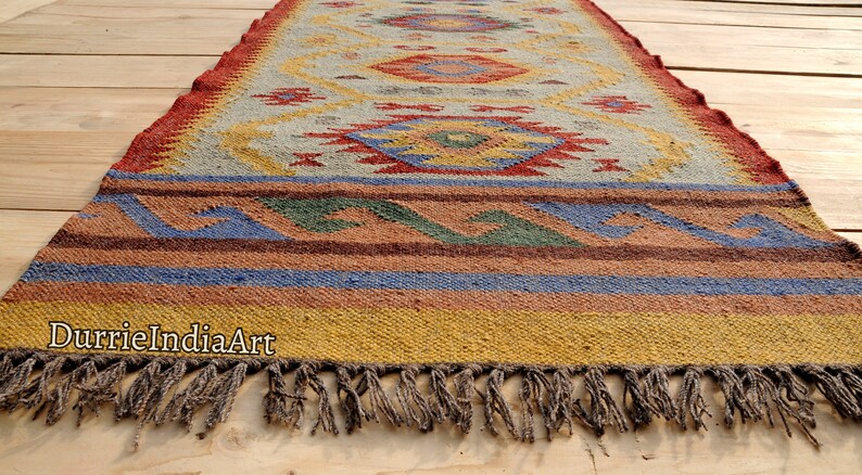 Pu&ograve; includere: Un tappeto colorato con motivo geometrico e frange. Il tappeto &egrave; realizzato in fibre naturali e ha un aspetto rustico e tessuto a mano.