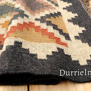 May include: A black, white, orange, and brown geometric patterned rug with fringe. The rug is folded and the fringe is visible. The rug is on a wooden floor.