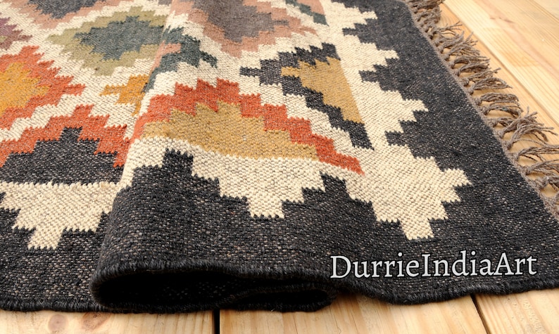 May include: A black, white, orange, and brown geometric patterned rug with fringe. The rug is folded and the fringe is visible. The rug is on a wooden floor.