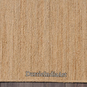 K&ouml;nnte beinhalten: Ein Naturfaser-Teppich aus Jute, einem nachhaltigen und strapazierf&auml;higen Material. Der Teppich hat eine gewebte Textur und eine hellbraune Farbe. Der Text "DurrieIndiaArt" ist in der rechten unteren Ecke des Teppichs sichtbar.