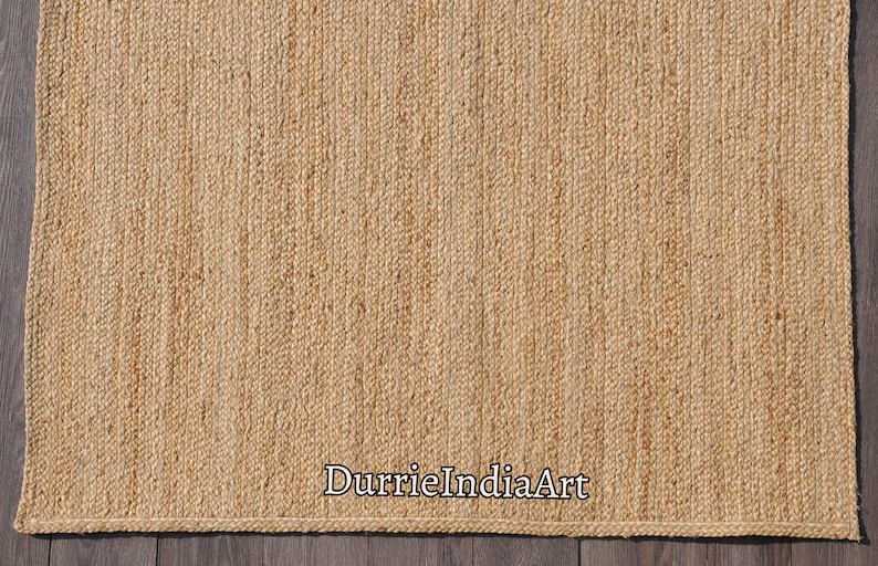 K&ouml;nnte beinhalten: Ein Naturfaser-Teppich aus Jute, einem nachhaltigen und strapazierf&auml;higen Material. Der Teppich hat eine gewebte Textur und eine hellbraune Farbe. Der Text "DurrieIndiaArt" ist in der rechten unteren Ecke des Teppichs sichtbar.