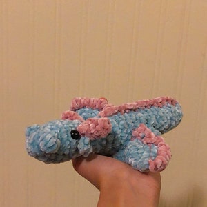 May include: A crocheted plush toy in the shape of a sea creature. The toy is light blue with pink accents and black eyes. The toy is being held by a hand wearing colorful bracelets.
