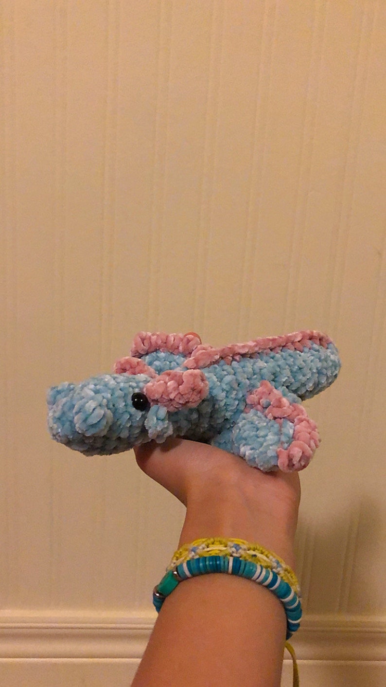 May include: A crocheted plush toy in the shape of a sea creature. The toy is light blue with pink accents and black eyes. The toy is being held by a hand wearing colorful bracelets.