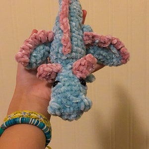 May include: A crocheted plush toy dragon with a light blue body and pink wings. The dragon has black eyes and a small, curled tail.