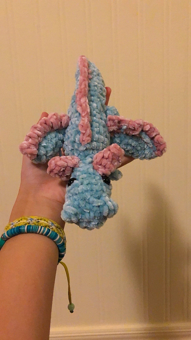 May include: A crocheted plush toy dragon with a light blue body and pink wings. The dragon has black eyes and a small, curled tail.