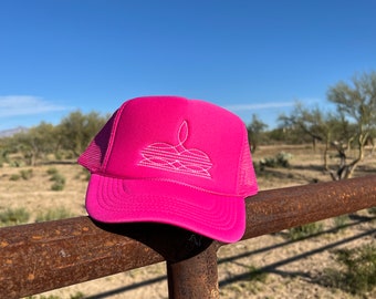 May include: A pink trucker hat with a white embroidered design on the front. The hat is sitting on a rusty metal fence.
